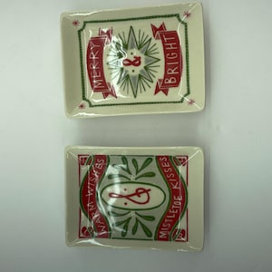 May include: Two rectangular ceramic trays with holiday designs. The top tray has the words "MERRY" and "BRIGHT" on red banners. The bottom tray has the words "WARM WISHES" and "MISTLETOE KISSES" on red banners. Both trays have green and red accents.