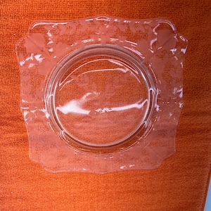 Puede incluir: Plato de vidrio transparente con forma cuadrada y bordes festoneados. El plato presenta un diseño floral grabado alrededor de los bordes y un centro circular. El plato está sobre una superficie naranja.