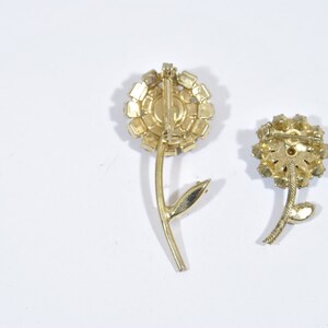 May include: Two gold-toned flower brooches with clear rhinestone accents. The brooches have a simple stem design.