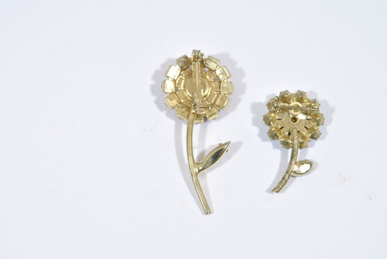 May include: Two gold-toned flower brooches with clear rhinestone accents. The brooches have a simple stem design.
