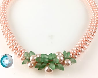 May include: A delicate necklace featuring a strand of light pink pearls. The necklace has a floral accent made of green leaves and larger pink pearls. The clasp is silver. The Ancient Moon logo is in the lower left corner.
