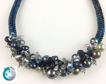 May include: A handcrafted necklace featuring a dark blue beaded collar. The necklace is adorned with clusters of dark blue, gray, and iridescent beads, pearls, and silver-toned metal accents. The necklace has a unique and elegant design.