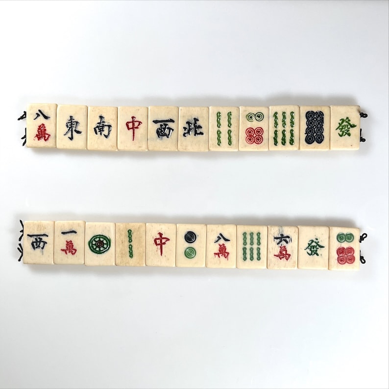 May include: Two rows of vintage Mahjong tiles, each with black, red, and green symbols and characters. The tiles are off-white with black string connecting them. The game pieces are arranged horizontally against a white background.