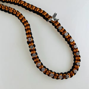 May include: A braided cord necklace featuring alternating black and copper-colored nylon strands. The cord is embellished with small, circular silver-toned flower-shaped metal beads and a single silver-toned hummingbird charm attached to the braid.