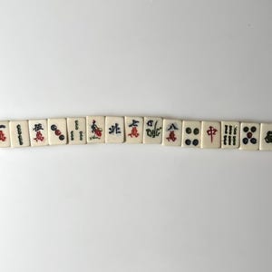May include: A row of off-white Mahjong tiles with black, red, and green symbols and characters. The tiles are arranged horizontally against a white background. The symbols include dots, bamboo, and Chinese characters.