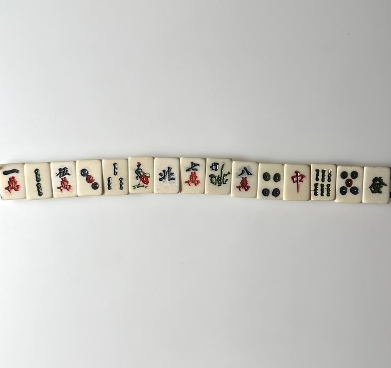 May include: A row of off-white Mahjong tiles with black, red, and green symbols and characters. The tiles are arranged horizontally against a white background. The symbols include dots, bamboo, and Chinese characters.