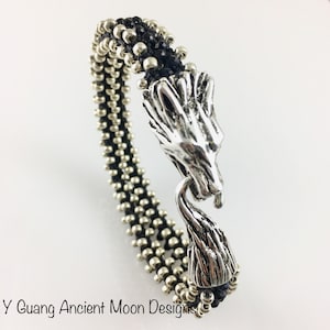 Könnte beinhalten: Ein silbernes Drachenkopf-Armband mit einem schwarzen Perlenband. Das Armband hat einen detaillierten Drachenkopfverschluss und besteht aus kleinen, runden silbernen Perlen. Der Text "Y Guang Ancient Moon Designs" befindet sich unten.