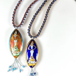 May include: Two oval pendant necklaces with braided cords. Each pendant features a painted figure in a dress. One pendant has an orange background, the other is blue. Both have beaded tassels and are on a white background.