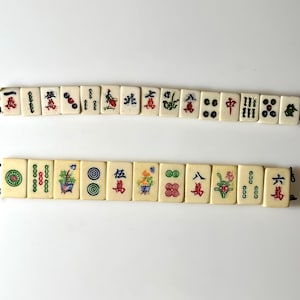 May include: Ivory-colored Mahjong tiles with red, green, and blue symbols and characters. The tiles are arranged in two rows, showcasing various designs including flowers, circles, and Chinese characters. The game pieces are displayed against a white background.