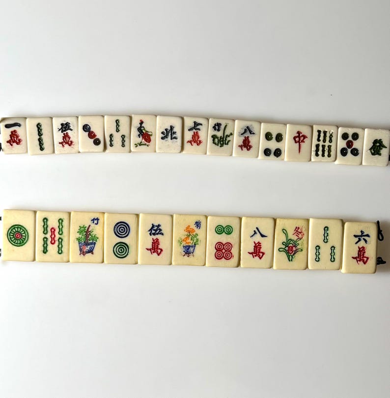 May include: Ivory-colored Mahjong tiles with red, green, and blue symbols and characters. The tiles are arranged in two rows, showcasing various designs including flowers, circles, and Chinese characters. The game pieces are displayed against a white background.