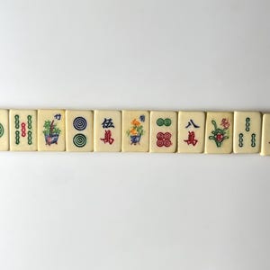 May include: A row of off-white Mahjong tiles with colorful symbols and Chinese characters. The tiles feature green, red, and blue designs, including flowers, circles, and bamboo. The tiles are arranged horizontally against a white background.
