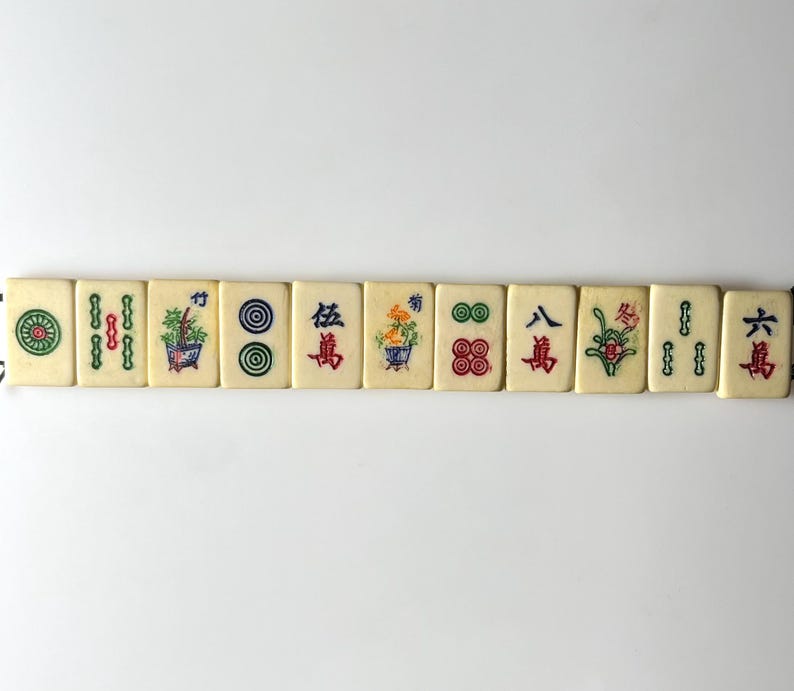 May include: A row of off-white Mahjong tiles with colorful symbols and Chinese characters. The tiles feature green, red, and blue designs, including flowers, circles, and bamboo. The tiles are arranged horizontally against a white background.