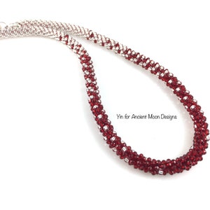 May include: A handmade necklace with alternating rows of deep red and silver beads. The beads are tightly woven, creating a textured, rope-like appearance. The necklace is displayed against a white background. Text: Yin for Ancient Moon Designs.
