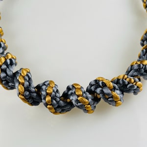 May include: A decorative cord featuring a twisted, spiral design constructed from interwoven strands of metallic gold and slate gray thread. The rope-like structure displays a consistent, repeating pattern of alternating colors throughout its length.