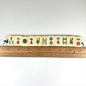 May include: A row of vintage ivory-colored Mahjong tiles with black string attached. The tiles feature red, green, and blue symbols and characters. A wooden ruler is placed below the tiles, showing measurements in inches.