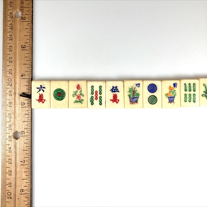 May include: A row of vintage ivory-colored Mahjong tiles with colorful illustrations and Chinese characters. The tiles feature various designs, including flowers, circles, and bamboo. A wooden ruler is in the image, showing inches.