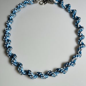 May include: A blue and silver twisted rope necklace with a silver clasp. The necklace is made of tightly woven blue and silver threads, creating a spiral pattern. The clasp features decorative floral accents.