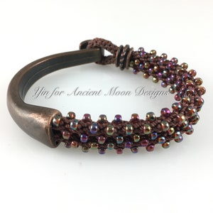 May include: A handcrafted bracelet featuring a dark brown woven cord adorned with iridescent beads. The bracelet has a bronze-colored metal clasp. The text "Yin for Ancient Moon Designs" is visible on the bracelet.