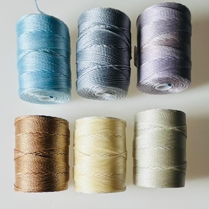 以下が含まれることがあります： 青、グレー、ベージュの色合いのカラフルな糸が6つのスプールに巻かれています。糸は小さなスプールにしっかりと巻かれており、わずかな光沢があります。色は水色からソフトベージュまであり、さまざまなクラフトプロジェクトに適しています。