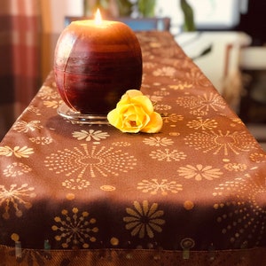 May include: A brown table runner with a gold and white geometric pattern. A brown candle with a yellow flame is on the runner, next to a yellow flower.