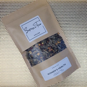 May include: A brown paper bag with a white label that says "Sereni Tea House" and a smaller white label that says "Premium Women's Health Herbal Tea". The bag is filled with loose leaf tea.