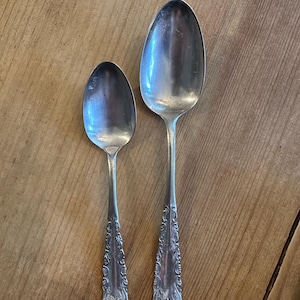 Puede incluir: Dos cucharas plateadas con detalles ornamentales en el mango sobre una superficie de madera. Una cuchara es más pequeña que la otra. Las cucharas tienen una superficie reflectante y los mangos presentan un diseño floral detallado.