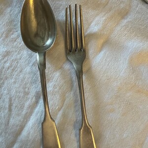 May include: A silver spoon and fork. The spoon has a rounded bowl and a long handle with a rectangular end. The fork has four tines and a matching handle. Both are made of shiny metal.