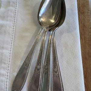 May include: Five antique silver spoons with detailed floral and scrollwork patterns on the handles. The spoons are displayed on a white linen cloth, highlighting their polished surfaces and vintage appeal. These spoons would be a classic addition to any dining set.