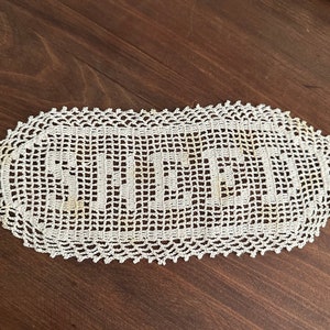 May include: Cream-colored, oval-shaped doily with the word "SWEETNESS" crocheted in the center. The doily has a scalloped edge and is placed on a dark brown wooden surface. A vintage-style home decor item.