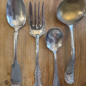 Può includere: Un assortimento di posate antiche, tra cui un cucchiaio grande, una forchetta, un cucchiaino e un mestolo. Le posate sono in metallo lucido color argento e presentano dettagli decorativi ornati. Gli utensili sono disposti su una superficie di legno.