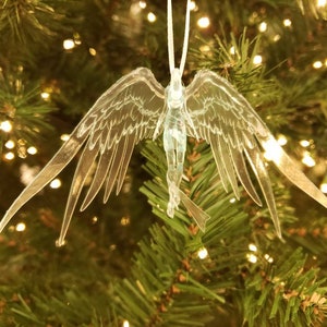May include: A clear, angel-shaped Christmas ornament with outstretched wings, hanging from a white string. The ornament is detailed with etched lines and is suspended in front of a Christmas tree with twinkling lights.