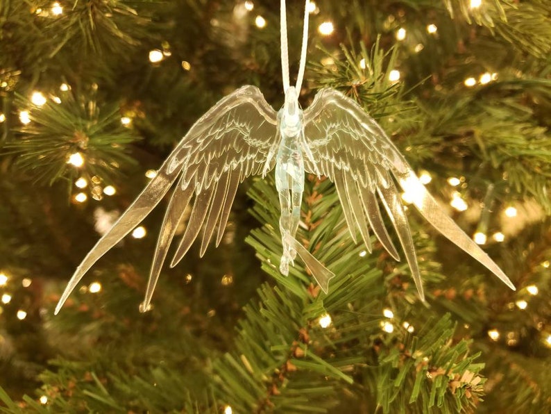May include: A clear, angel-shaped Christmas ornament with outstretched wings, hanging from a white string. The ornament is detailed with etched lines and is suspended in front of a Christmas tree with twinkling lights.