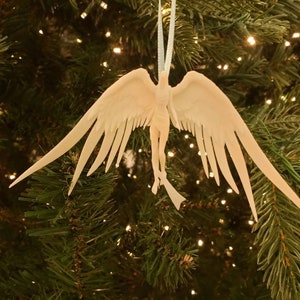 May include: A white angel Christmas ornament with outstretched wings hangs from a white string against a backdrop of a decorated Christmas tree. The angel has detailed feathering and a flowing form.