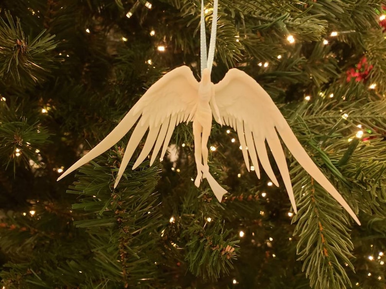 May include: A white angel Christmas ornament with outstretched wings hangs from a white string against a backdrop of a decorated Christmas tree. The angel has detailed feathering and a flowing form.