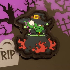 May include: A Halloween-themed patch featuring a cartoon witch stirring a bubbling cauldron. The witch is green with a black hat and is set against a black and red background. A tombstone with "RIP" is also visible.