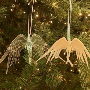 May include: Two angel-shaped Christmas ornaments hanging on a green Christmas tree. One is clear with a pink ribbon, the other is beige with a light blue ribbon. Both have outstretched wings and are detailed.