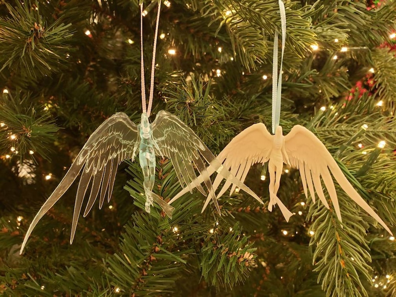 May include: Two angel-shaped Christmas ornaments hanging on a green Christmas tree. One is clear with a pink ribbon, the other is beige with a light blue ribbon. Both have outstretched wings and are detailed.