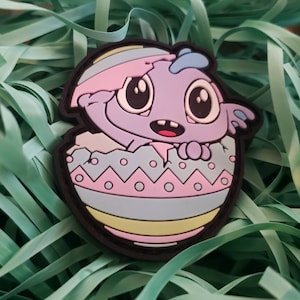 May include: A purple and blue cartoon monster peeking out of a pastel pink, blue, and yellow Easter egg. The egg has a cracked opening and a black outline.