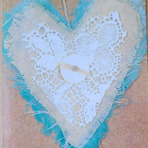 May include: A heart-shaped piece of fabric with a lace overlay and a frayed edge. The fabric is a light blue color and the lace is white. The heart is on a brown background.