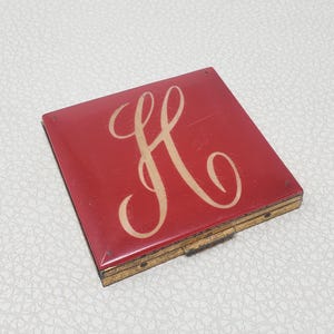 Op de afbeelding: Een rode compact met een goudkleurig frame. De compact heeft een grote, cursieve letter "H" in een crème kleur op de bovenkant. De compact is vierkant en lijkt vintage te zijn.