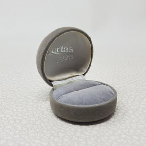 Puede incluir: Una caja de joyería gris cubierta de terciopelo, abierta para revelar un interior gris con dos ranuras para anillos. El interior de la tapa es gris claro con el texto "Luria's Est. 1898". La caja es redonda con tapa abisagrada, sobre fondo blanco.