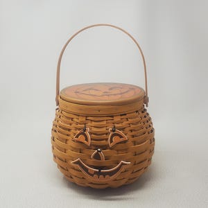 May include: A round, woven brown basket with a matching lid featuring a pumpkin face design. The basket body has a carved wooden jack-o'-lantern face attached to the front, complete with eyes, a nose, and a smiling mouth. It has a thin metal handle.