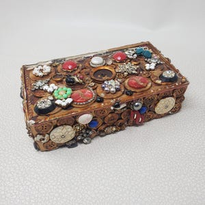 May include: A rectangular wooden box decorated with various buttons, beads, and embellishments. The top is adorned with red, white, and black decorative elements. The sides are covered in brown buttons.