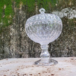 May include: A clear, footed glass candy dish with a lid. The dish has a round bowl with a textured, patterned design and a pedestal base. The lid has a small knob. The dish is sitting on a white lace tablecloth.