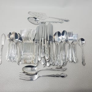 May include: A collection of shiny silver flatware, including knives, forks, spoons, and serving utensils. The silverware is arranged on a light-colored surface, showcasing a complete set for dining. The utensils have decorative handles.