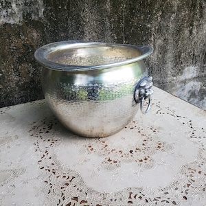 May include: A silver-colored metal bowl with a hammered texture and a wide, smooth rim. The bowl features a decorative lion head handle on the side, which includes a circular ring attachment. The surface of the metal reflects light across its curved body.
