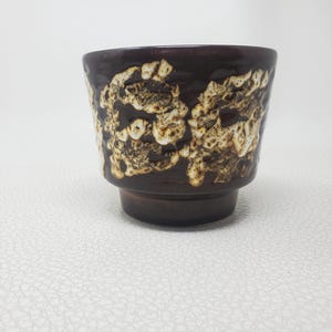 May include: A dark brown ceramic cup featuring a textured, raised cream-colored glaze pattern around the upper exterior. The vessel has a wide, flared rim that tapers down to a narrower, solid dark brown base, creating a distinct stepped profile.