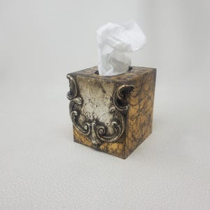 May include: A square tissue box cover featuring a faux marble finish in shades of gold and brown. The front panel is adorned with a prominent, raised silver-toned decorative scrollwork element. A single white tissue is pulled through the top opening.