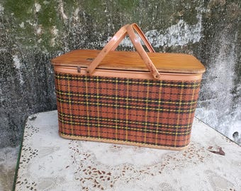 May include: A vintage picnic basket with a wooden lid and handle. The basket's body features a plaid pattern in shades of brown, black, and yellow. The handle is made of wood. The basket is sitting on a white lace tablecloth.