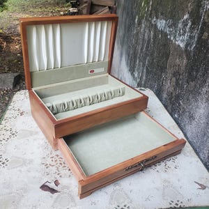 Puede incluir: Un joyero de madera con tapa abatible y un cajón extraíble. El interior cuenta con un forro de tela blanca plisada en la tapa, un compartimento forrado de fieltro verde claro con ranuras y un cajón abierto a juego en la parte inferior.